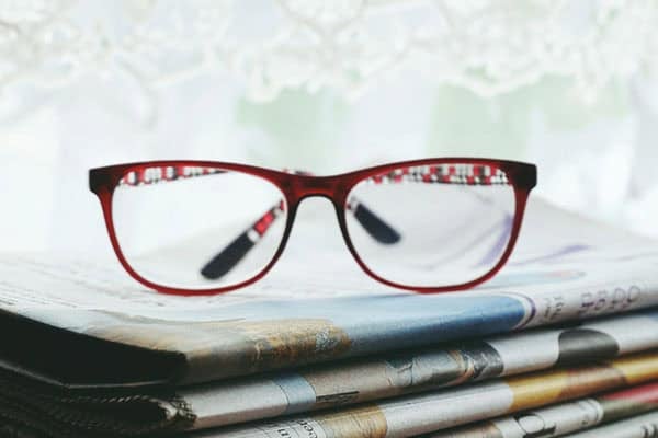 Stack of local newspapers in front of a window with a pair of reading glasses resting on top